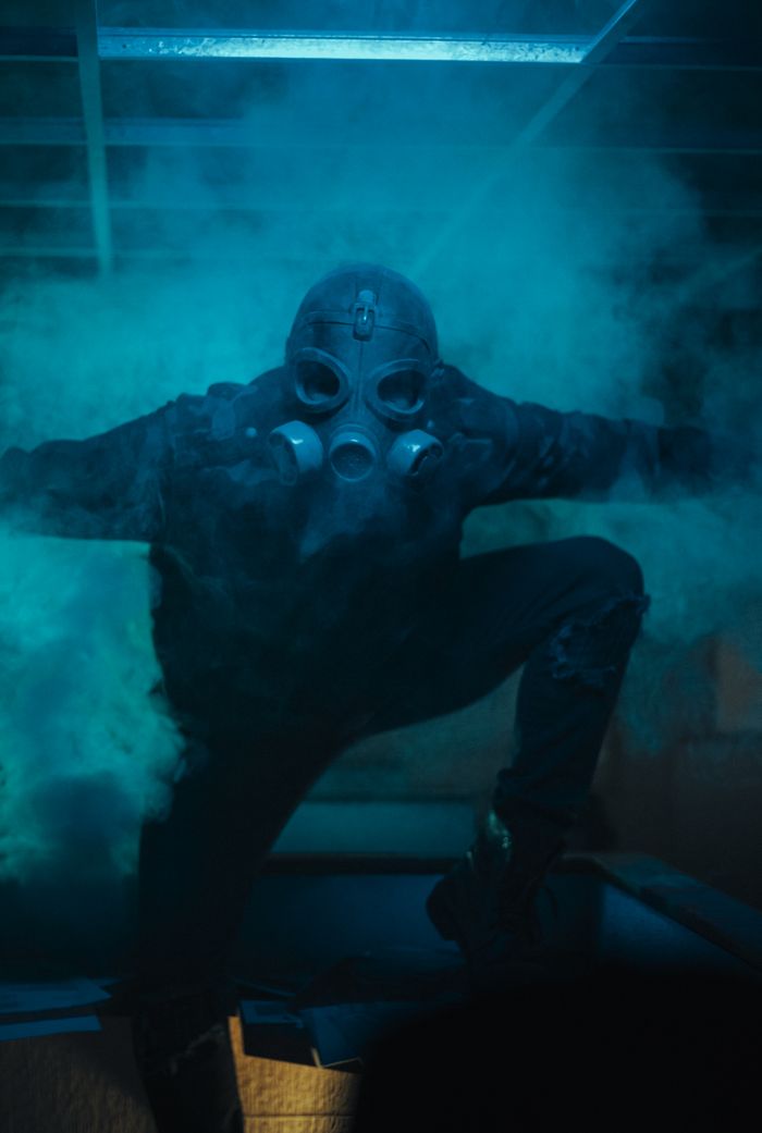 A haunting figure in a gas mask poses dramatically in a blue-lit smoky room.