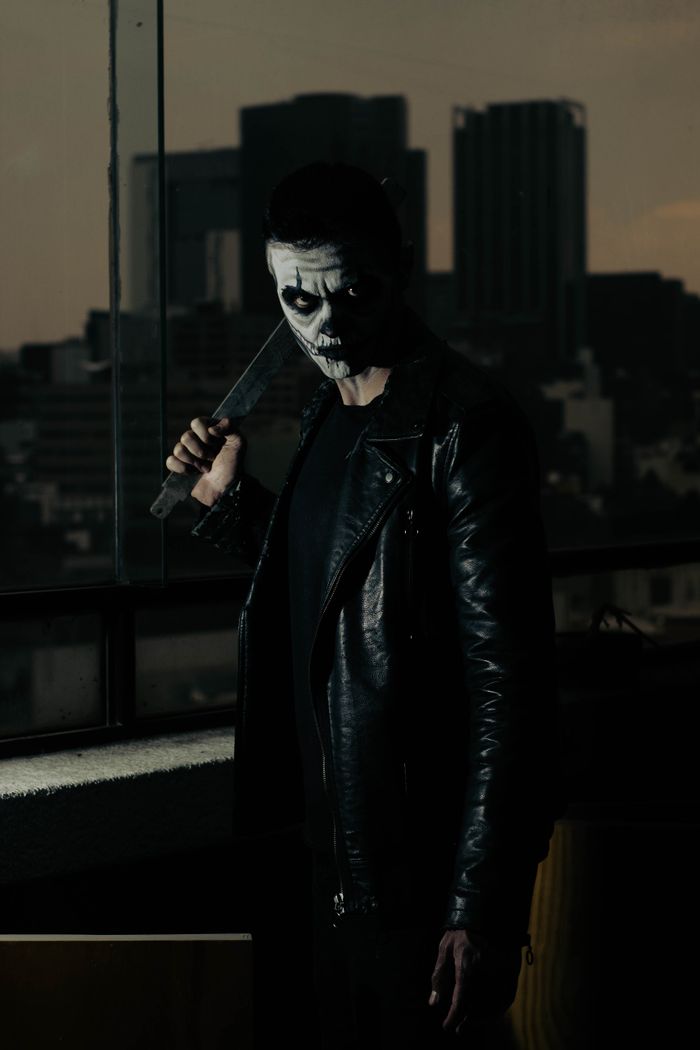 A man in a leather jacket and horror-themed mask holds a knife on a rooftop at dusk.