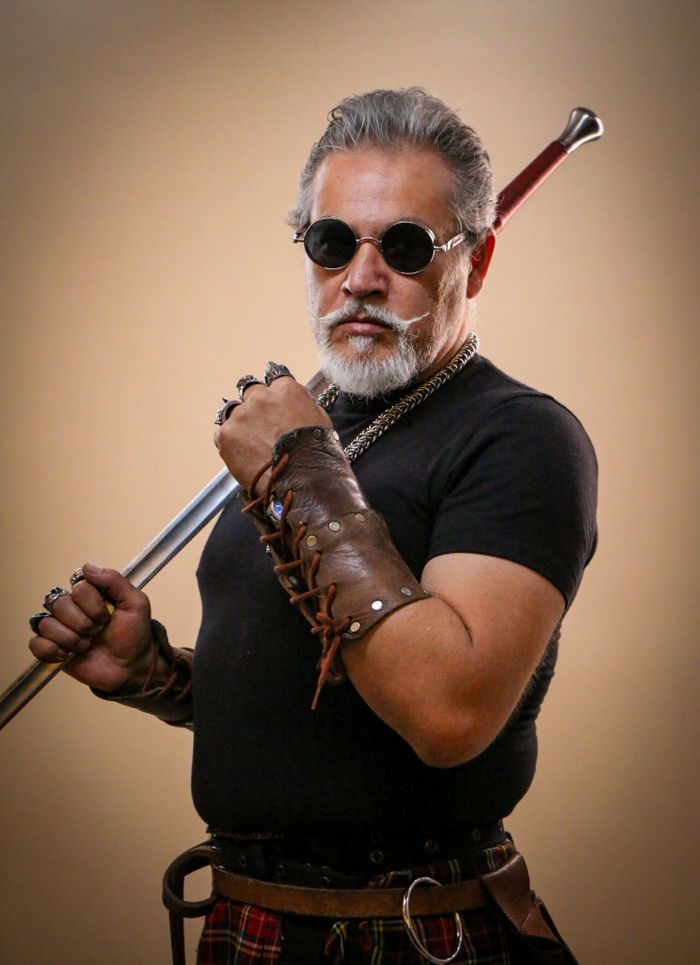 Confident mature man with beard, wearing leather cuffs, holding a sword against a neutral background.